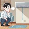 An illustration of a Japanese person troubled by an air conditioner leaking water. The person looks concerned, staring at a puddle forming beneath the AC unit. The setting is indoors, with a modern living room background. The illustration style is clean and simple, with soft colors and expressive facial features. Aspect ratio: 16:9.